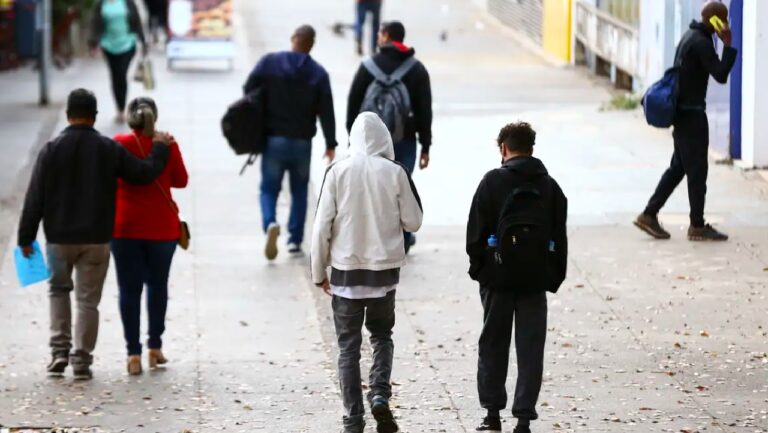 Pessoas caminhando em calçada urbana em dia frio com casacos e mochilas seguindo em direções diferentes