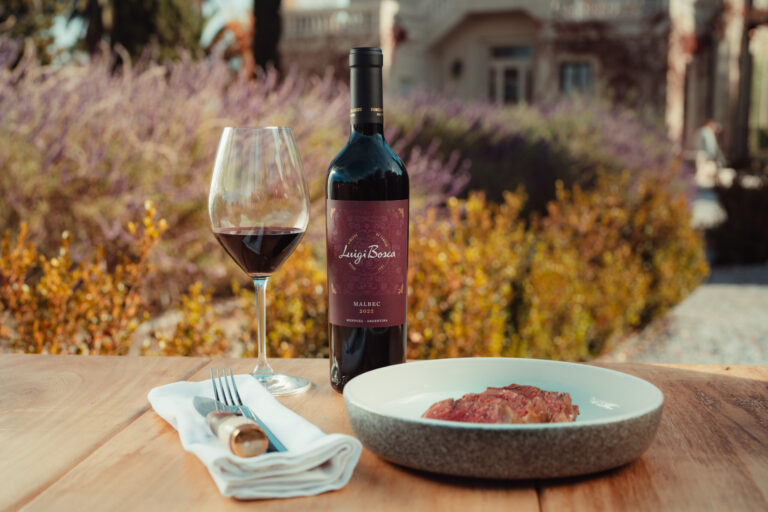 Garrafa de Luigi Bosca Malbec 2022 com taça de vinho e prato com carne em mesa ao ar livre em cenário de vinícola