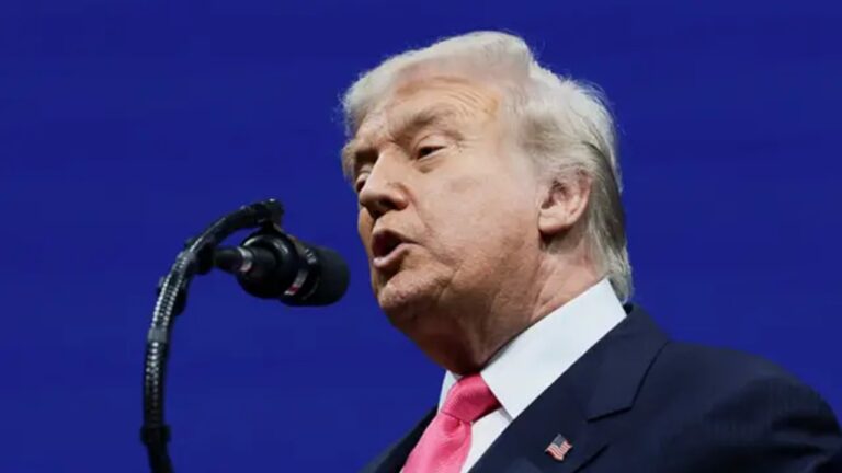 Homem de terno escuro e gravata rosa falando ao microfone em palco com fundo azul