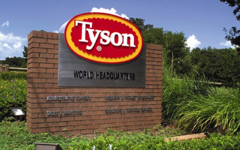 Placa de entrada da sede mundial da Tyson com logotipo vermelho e amarelo em estrutura de tijolos cercada por vegetação
