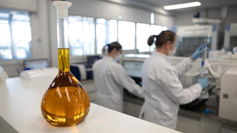 Frasco volumétrico com líquido âmbar em bancada enquanto técnicas operam equipamentos em laboratório de análises químicas