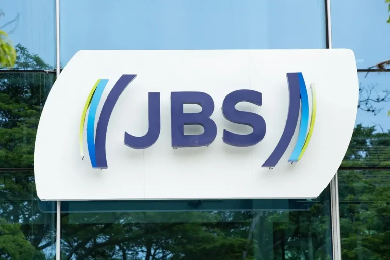 Logotipo da JBS em painel externo de prédio com fundo envidraçado e árvores refletidas