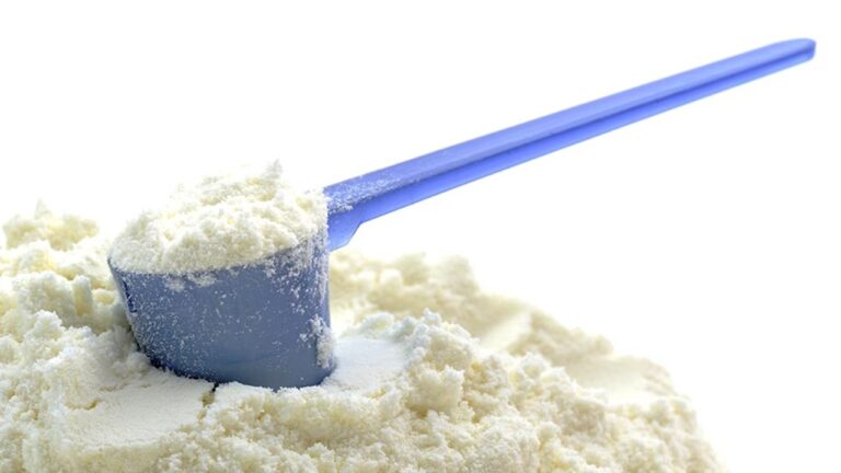 Medidor azul com suplemento em pó branco apoiado sobre monte do produto em fundo branco