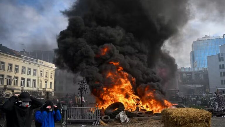 Manifestantes observam barricada em chamas com pneus queimando e fumaça preta em área urbana cercada por prédios