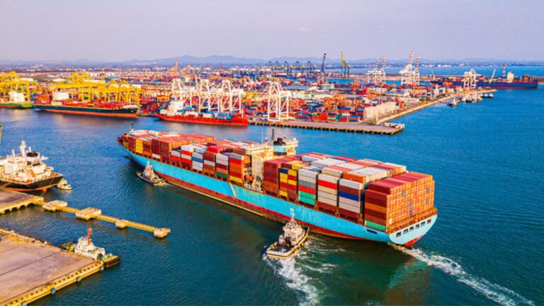 Navio cargueiro com contêineres navega em porto com guindastes e terminal ao fundo durante operação de comércio exterior