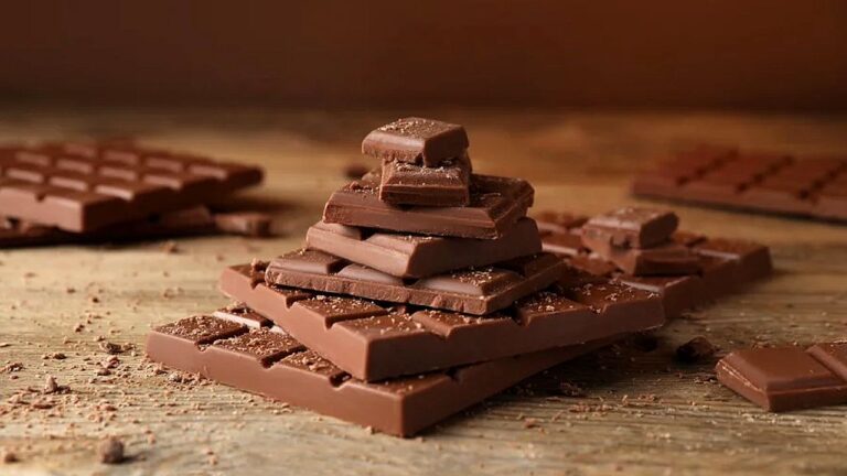 Tabletes de chocolate empilhados sobre mesa com pedaços ao redor em imagem sobre consumo e indústria do chocolate