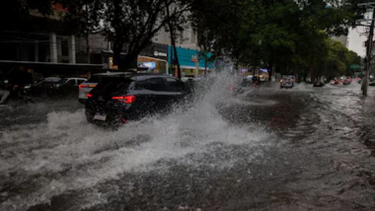 Carro atravessa rua alagada durante chuva forte em área urbana em imagem sobre enchentes e transtornos no trânsito