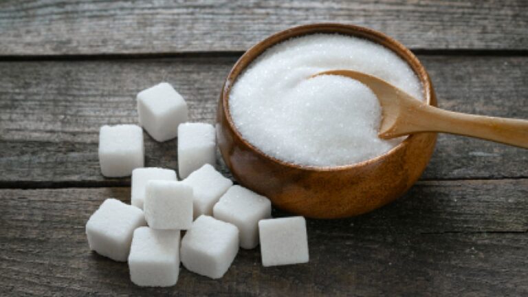 Açúcar refinado em tigela com colher de madeira e cubos de açúcar sobre superfície rústica ilustrando consumo do produto