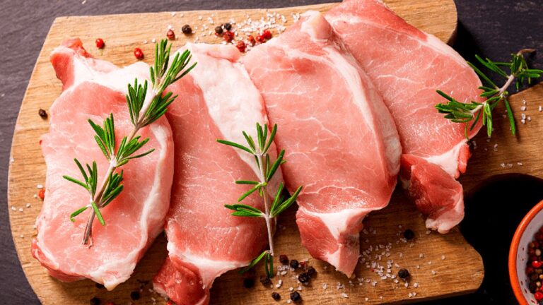 Cortes de carne suína crua dispostos em tábua com ramos de alecrim e grãos de pimenta em preparação culinária