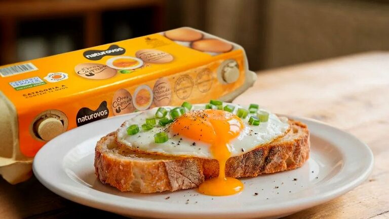 Fatia de pão com ovo frito de gema mole em prato com caixa de ovos ao fundo ilustrando consumo de alimento básico