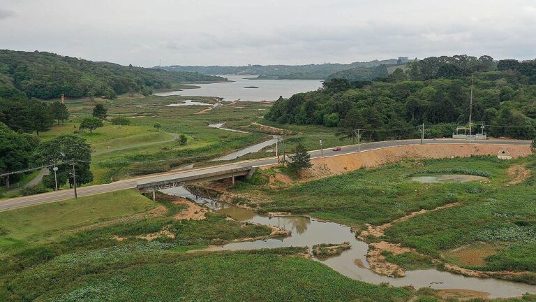 Represa com nível baixo de água cercada por vegetação e estrada em área de seca ambiental