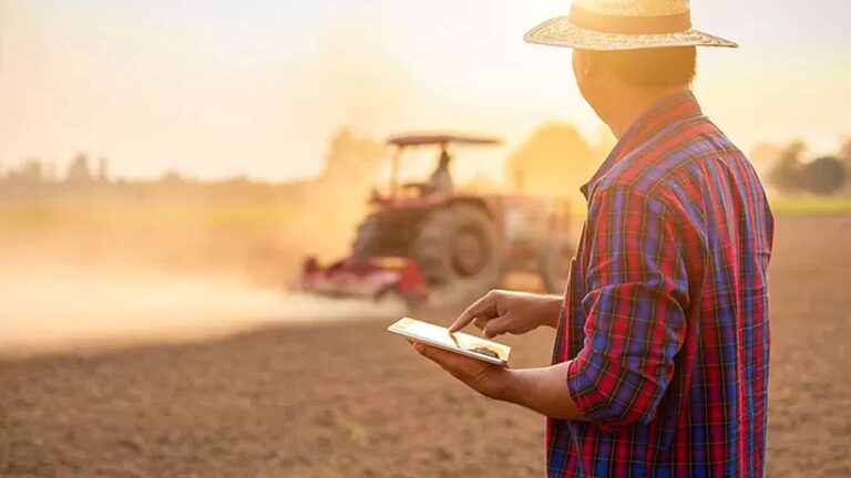 Produtor rural usa tablet para monitorar lavoura enquanto trator trabalha no campo durante atividade agrícola