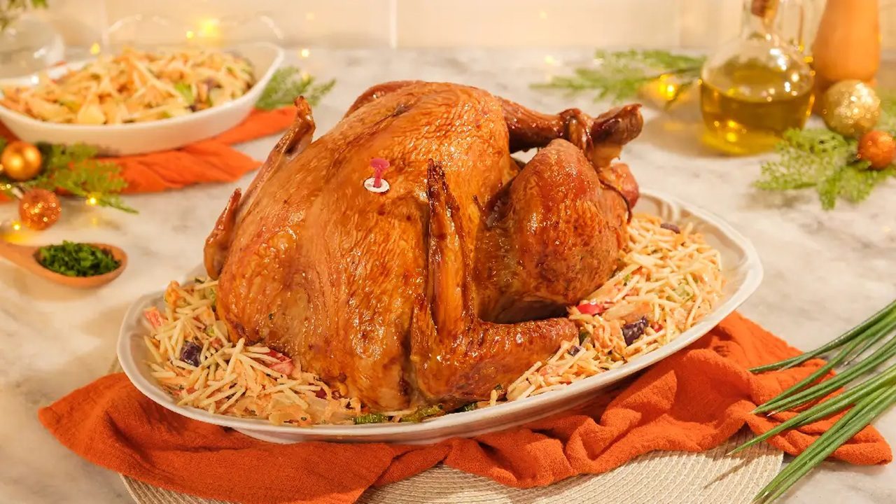 Peru assado inteiro sobre salpicão em travessa branca com decoração natalina ao fundo